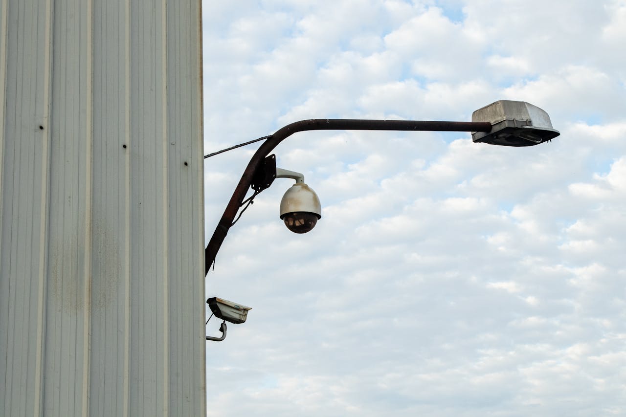 Street lamp and CCTV camera mounted on a building against a cloudy sky.